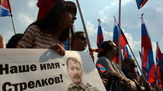 Fotografia colorida mostra gruposportbettvmanifestantes com bandeiras da Rússia e um cartaz com a imagemsportbettvum homem brancosportbettvbarba