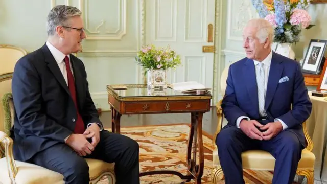 Premier Wielkiej Brytanii Keir Starmer spotyka się z królem Karolem w Pałacu Buckingham po raz pierwszy po wygraniu wyborów powszechnych w lipcu 2024 r. Obaj siedzą na krzesłach i ubrani są w ciemne garnitury, białe koszule i krawaty.