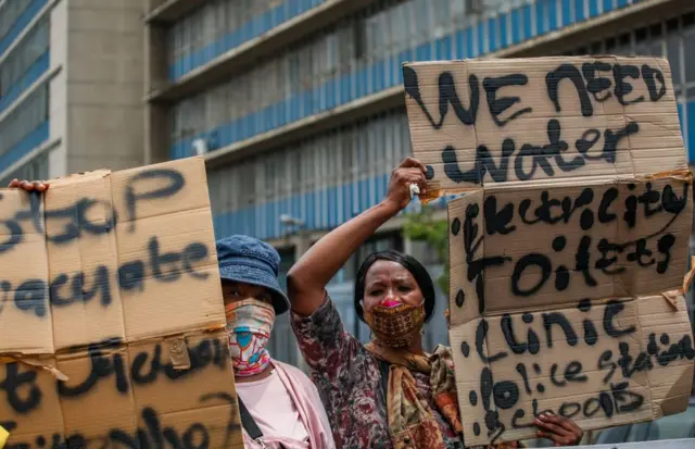 Two women hold up signs demanding access to amenities and services, including water and toilets.
