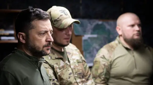 Зеленський під час візиту на Харківщину в серпні - стоять троє людей в темному приміщенні, зосереджений президент і двоє військових
