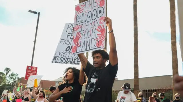 Fotografia colorida mostra um homem e uma mulher negros protestando com cartazes e diversas pessoas brancas protestandobetboo 793segundo plano