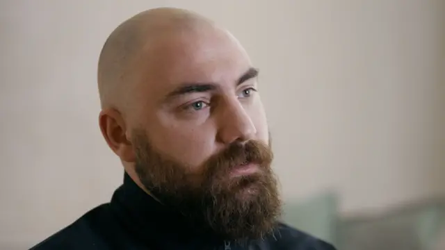 У Костянтина поголена голова, рудувата борода та світлі очі. Він одягнений у чорну водолазку