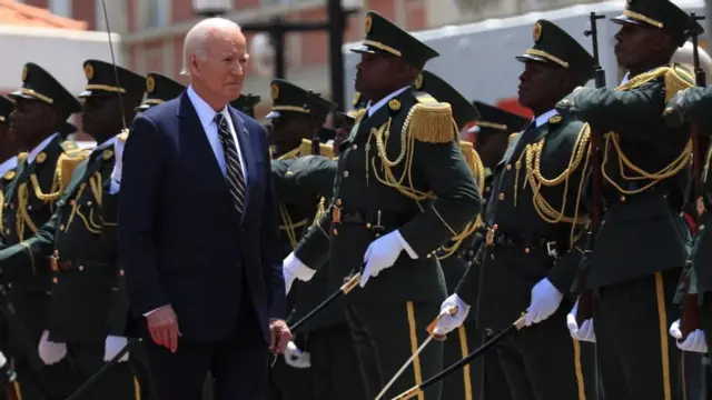 Joe Biden passe en revue une garde d'honneur à son arrivée au palais présidentiel de Luanda. Il longe un groupe de soldats en uniforme de cérémonie, gantés de blanc et brandissant des épées.