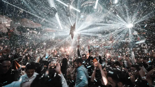 Masquerade Gece Kulübü'ndeki ana dans pistinde yüzlerce kişi dans ediyor. Işık oyunları yapılıyor ve konfeti atılıyor. Bazı kişiler ortamı telefonlarıyla kayıt altına alıyor. 
