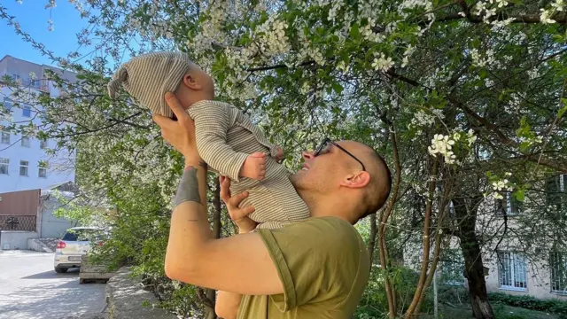 Давид Чичкан підіймає догори свого сина на фоні яблуневого дерева