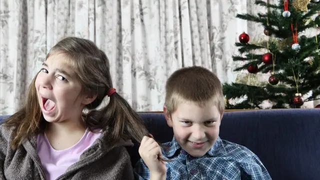 Siblings squabbling at Christmas: a boy and a girl sitting on a blue sofa, the boy is pulling the girl's pigtail. Christmas tree in the background