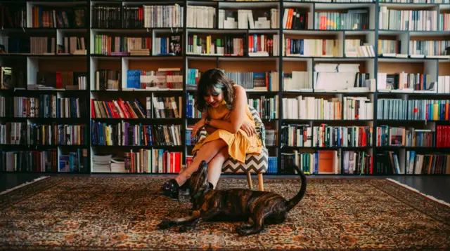 Devojka sedi na stolici u biblioteci, gleda u psa koji se igra na podu