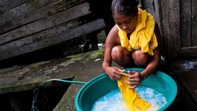 Una mujer lava ropa frente a una casa de madera.