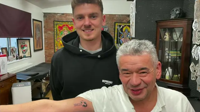 Un hombre con una sudadera con capucha negra está detrás de un hombre con un polo blanco que tiene un pequeño balón de fútbol con "SH22" tatuado en su bíceps.