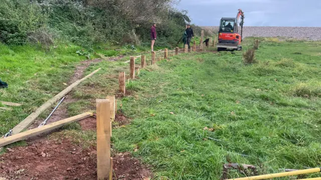 Dos personas y una excavadora trabajan en la construcción del paseo marítimo en un campo. Hay una hilera de postes de madera en el suelo y se ve un banco de guijarros al fondo.