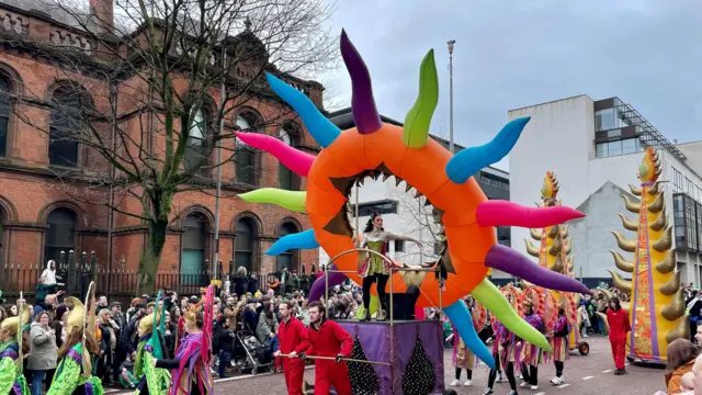 Di image show a vibrant parade wit various participants in colourful costumes. Di central focus na large, inflatable structure wey resemble a sun wit multicoloured rays, mounted on a purple platform. A performer stand inside di structure, dressed in a green and gold outfit. Surrounding di structure na participants in red outfits pushing di platform, and odas dey in green and purple costumes holding decorative staffs. Di parade dey take place on a street lined wit spectators, wit a historic brick building and a modern building in di background. Di scene dey lively and festive, indicating a celebration or festival.