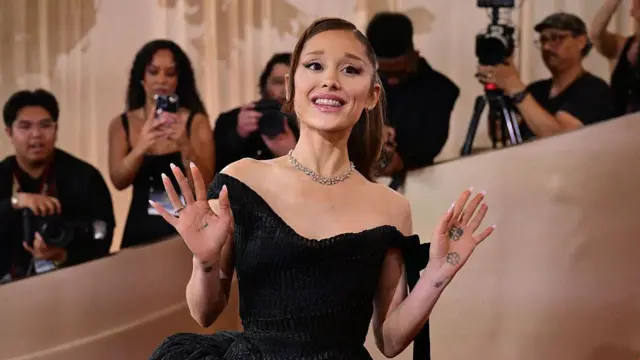 Siyah bir elbise giymiş Ariana Grande iki elini de havaya kaldırmış ve hayranlarını selamlıyor. Arka tarafında çok sayıda gazeteci de onun fotoğrafını çekiyor