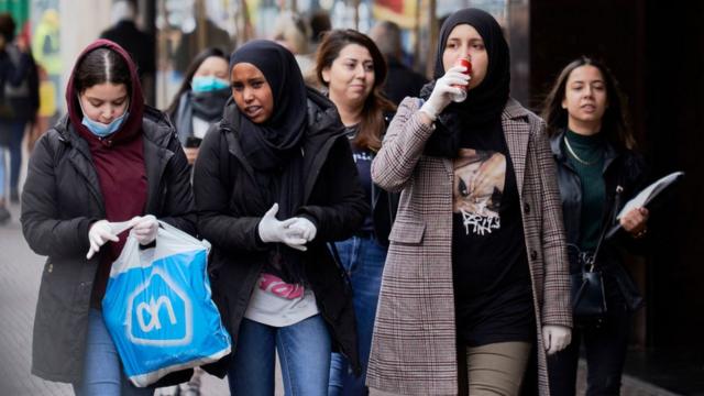 Um grupo de jovens mulheres com lenços de cabeça andando na rua na Holanda. Algumas usam máscaras, outras usam luvas e outras não usam qualquer proteção