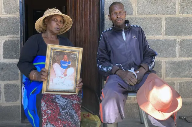 Mulher segura retrato do filho; ao lado dela, um homem adulto