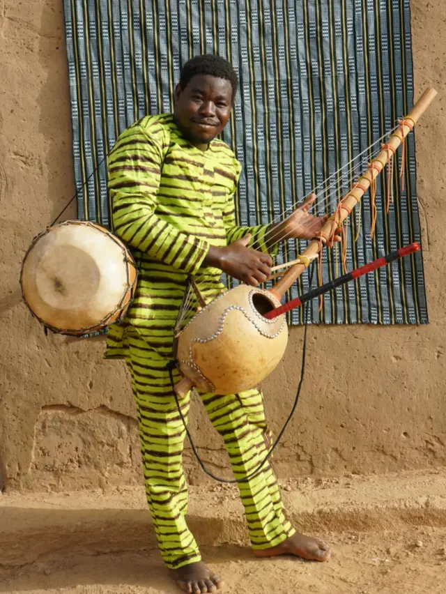 Maboudou Sanou pose avec un tambour bendré, un instrument ressemblant à une harpe appelé kora et sa flûte - les trois instruments qu'il joue dans l'opéra de Fujiie. Comme beaucoup de musiciens issus de familles de griots, et dans le groupe lui-même, Sanou joue de plusieurs instruments, se décrivant lui-même comme "polyvalent".