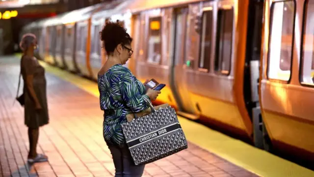 승객들이 지하철 승강장에 서 있다. 가까이 보이는 한 사람은 스마트폰 화면을 보고 있다