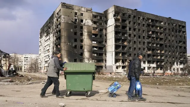two men push a wheelie bin past bombed flats