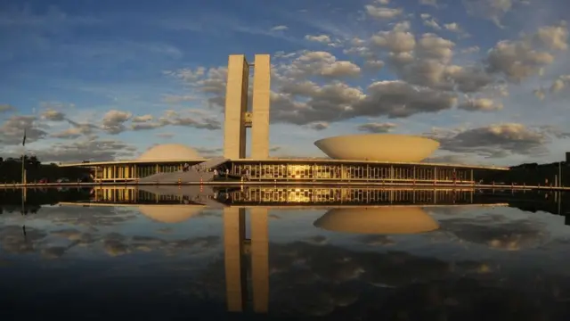 Prédio do Congresso refletido num espelho d'água sob um céu azul com nuvens