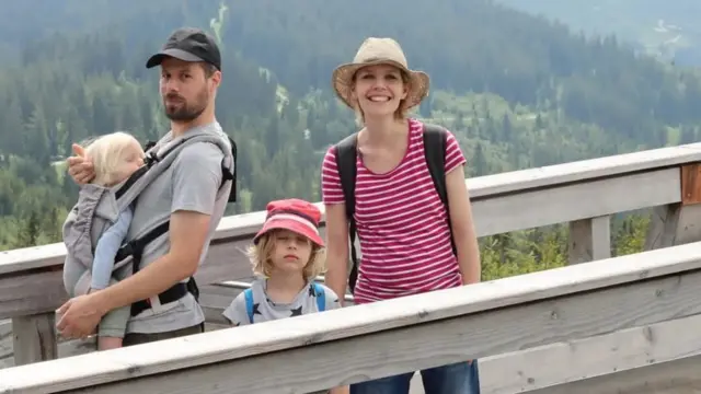 Um homem branco carrega um bebe loiro no colo, ao seu lado uma mulher e uma crianca brancas sorriem para a camera