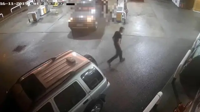 На записі з камери спостереження видно, як людина йде мокрим майданчиком біля автозаправки вночі. Поруч припарковані кілька автомобілів біля паливних колонок, у верхньому лівому куті видно позначку часу.