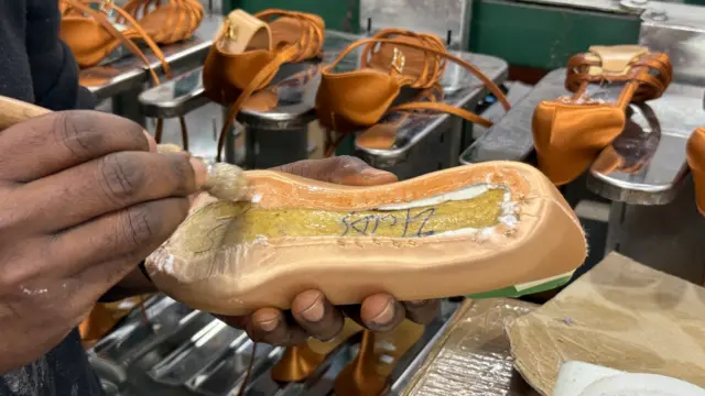 Primer plano de la fabricación de un zapato. Se sostiene un zapato en una mano mientras con la otra se aplica pegamento a la suela con un pincel. Al fondo se ven filas de zapatos.