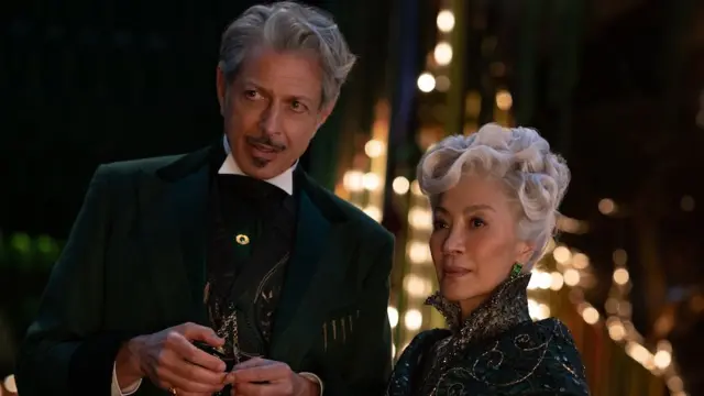 Jeff Goldblum veste um terno escuro e Michelle Yeoh está com um top enfeitado. Ambos têm olhares curiosos em seus rostos