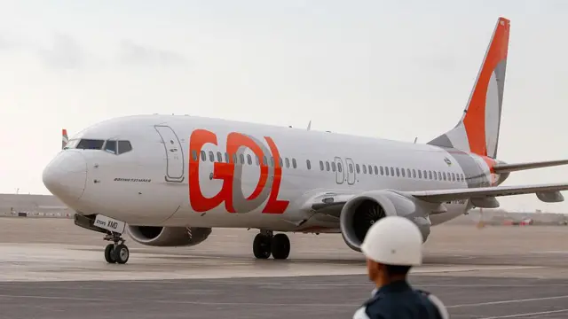 Avião da Gol estacionado no pátio de um aeroporto, enquanto um funcionário usando capacete observa a aeronave em primeiro plano.