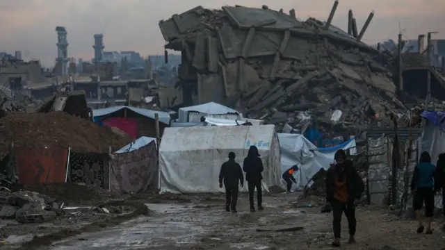 يسير فلسطينيون نازحون داخلياً بين أنقاض المباني المدمرة في حي الزيتون شرقي مدينة غزة، في يوم ممطر، بتاريخ 12 ديسمبر/كانون الأول 2025، وسط وقف إطلاق النار بين إسرائيل وحماس.