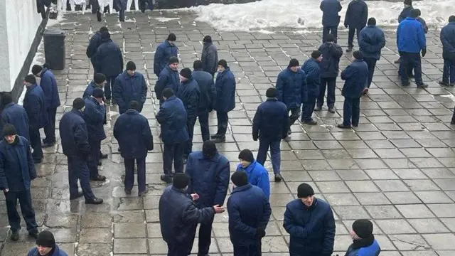 На плацу тюрми близько сорока чоловік у темно-синіх куртках і штанах, а також чорних шапках прогулюються. Вони роззосереджені невеликими групами по чотири-шість чоловік