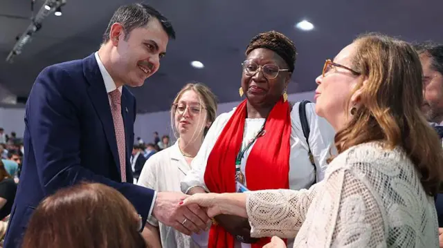 Türk bakan takım elbiseli karşısındaki kadının gözlerinin içine bakıyor, yanında Afrika kökenli gözlüklü bir kadın var.