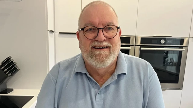 Ifoto y'umusaza wambaye umupira w'ubururu bwerurutse wambaye n'indorerwamo z'umukara, afite akanyamuneza mu maso. 