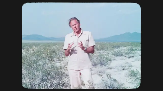 Attenborough de pie en un paisaje desértico, gesticulando frente a matorrales secos y montañas al fondo.