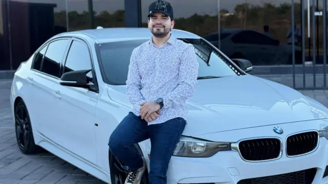 César Gastélum sentado sobre un BMW blanco. Lleva una gorra negra