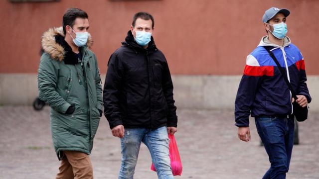 Três homens com máscaras e roupas de frio caminham em rua