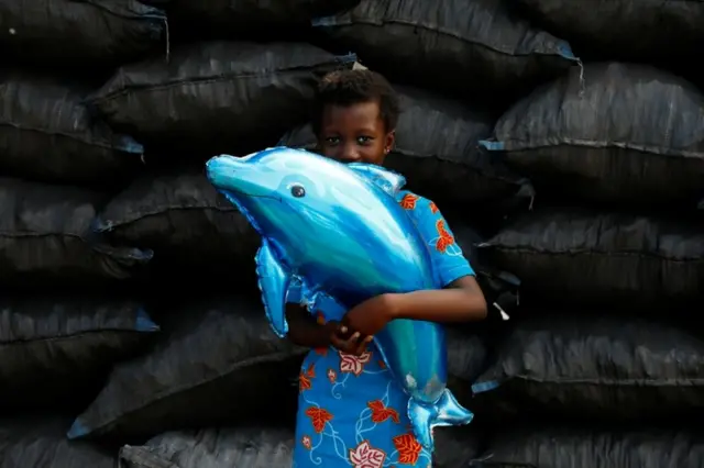 Wata yarinya sanye da kaya shuɗaye cikin wani murmushi mai haɗe da kunya ta riƙe kifin roba.