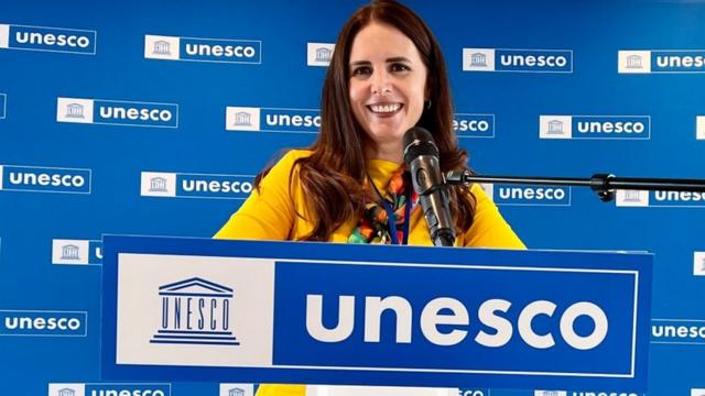 Elisa Guerra sorrindow malinowski pokerfrente a painel com logo da Unesco e pertow malinowski pokermicrofone