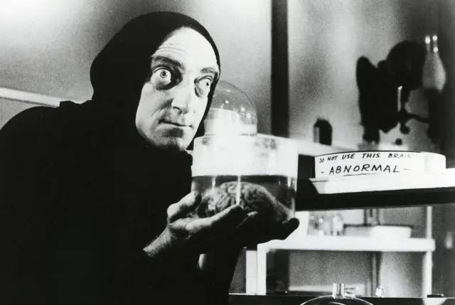 Чоловік у чорному з очима на викоті тримає склянку з мозком