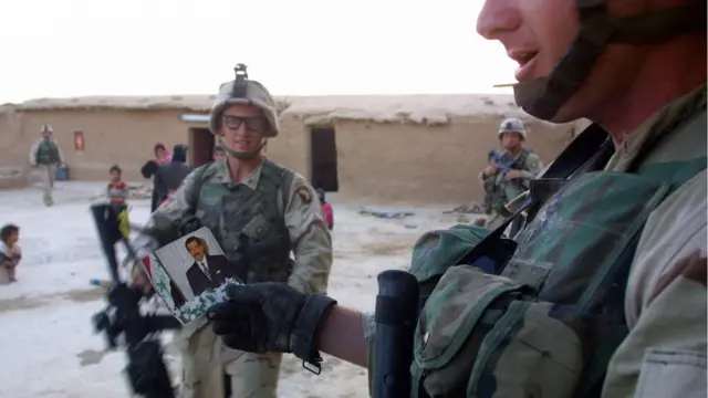 ইরাকী নেতা সাদ্দাম হুসেনের ছবি হাতে আমেরিকান সৈন্যরা আলাইবা গ্রামে বিভিন্ন বাড়িতে ক্ষমতাচ্যুত নেতাকে খুঁজছে