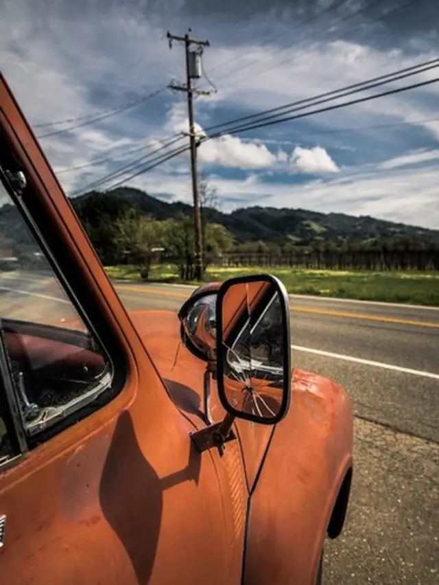Classic orange old truck with broken side mirror, with road, telephone pole and green hills behind it. Blue sky, midday.