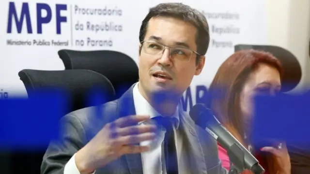 Deltan Dallagnol fala ao microfone, perto de painéis com logo do MPF