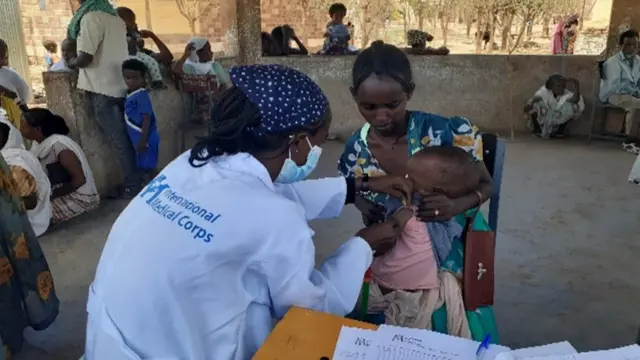 የኢንተርናሽናል ሜዲካል ኮርን አርማ የያዘ ነጭ ጋውን የለበሰች የጤና ባለሙያ ህጻን ልጅ እየከተበች