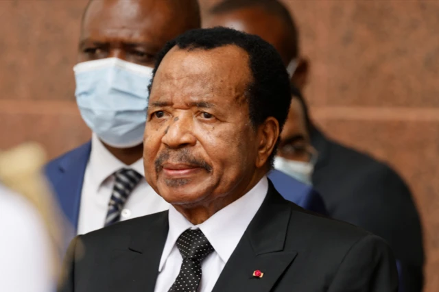 Rais wa Cameroon Paul Biya akiwa amevalia suti nyeusi, shati jeupe na tai yenye madoadoa meusi. Mwanamume kuna mwanamume aliyevalia barakoa