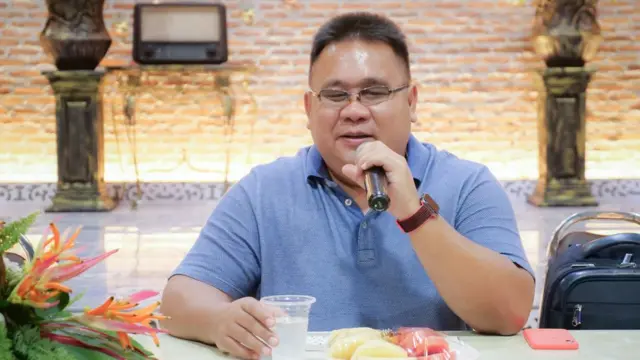 ภาพนายต่อพงศ์สวมเสื้อสีฟ้า มือซ้ายจับไมค์ยกขึ้นพูด