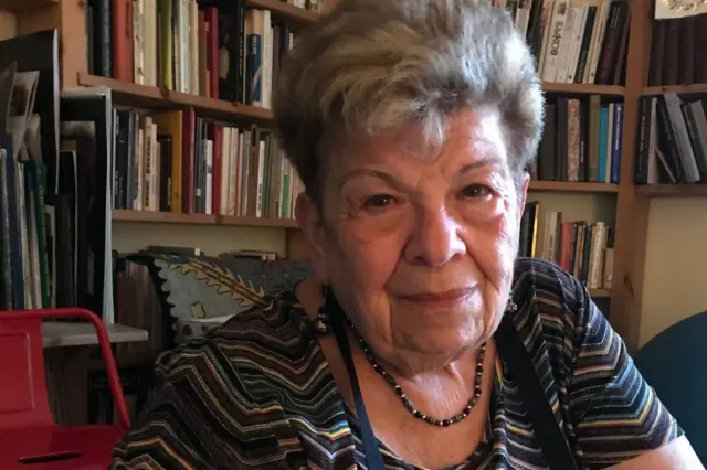 Наталья Рубинштейн, пожилая женщина в полосатой кофточке, сидит на фоне полок с книгами