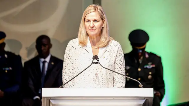 Kirsty Coventry, présidente du Comité international olympique, apparaît vêtue d'un chemisier blanc à manches longues, prononçant un discours depuis un pupitre blanc où deux microphones fins se rejoignent devant elle. L'arrière-plan, légèrement flou, laisse apparaître plusieurs hommes en costumes sombres et uniformes de style militaire, sous une lumière chaude beige et verte.