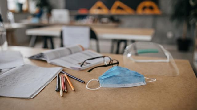 Em uma mesa, óculos de aluno, livros abertos, um protetor facial e uma máscara azul