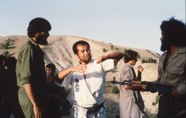 کوشیرو در حالی که لباس رزمی به تن دارد در حال آموزش حرکتی برای دو نفر از مجاهدین است