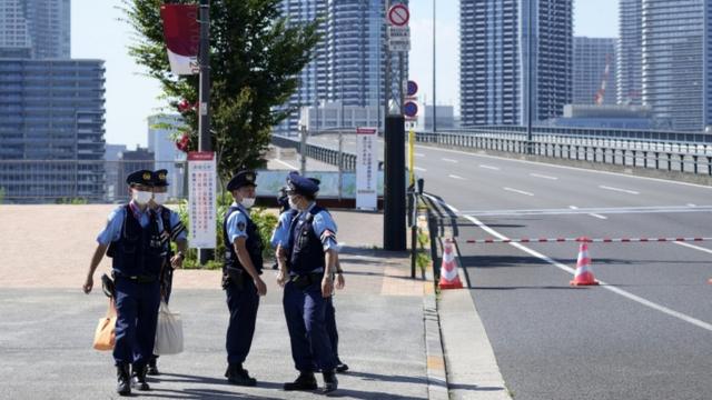 Policiais de máscara em frente a via de acesso e a prédios da vila olímpica