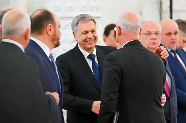 Кілька чоловіків у ділових костюмах стоять у колі та спілкуються між собою. Сцена виглядає як офіційний прийом або дипломатична зустріч у світлій урочистій залі з декоративною колоною на тлі.