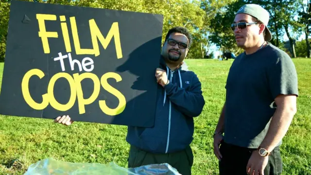 Dennis Flores, um homem latino baixobônus de apostas esportivas e cassino betanoóculos, segura um cartaz escrito 'filme a polícia',bônus de apostas esportivas e cassino betanoinglês, no meiobônus de apostas esportivas e cassino betanoum parque